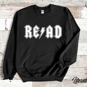 Puede incluir: Sudadera negra con un gr&aacute;fico blanco que dice "READ" en una fuente de estilo rock and roll con un rayo.