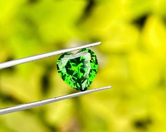 Op de afbeelding: Een hartvormige groene edelsteen met een facetgeslepen vorm, vastgehouden door een pincet tegen een wazige groene achtergrond.