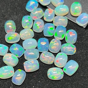Könnte beinhalten: Eine Sammlung von 25 losen, weißen Opal-Cabochons mit irisierenden Regenbogen-Blitzen. Die Opale sind oval geformt und haben eine glatte, polierte Oberfläche.