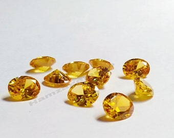 May include: A group of ten round, faceted, yellow gemstones. The gemstones are scattered on a white surface.