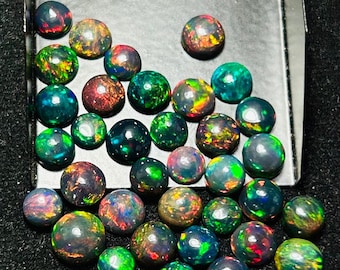 May include: A collection of round, iridescent black opals with green, red, and blue flashes. The opals are arranged in a loose pile on a black surface.