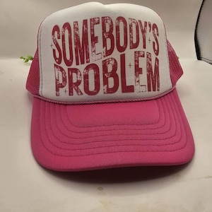 May include: A pink trucker hat with a white front panel. The text "SOMEBODY'S PROBLEM" is printed in red across the front. The hat has a pink brim and mesh backing.