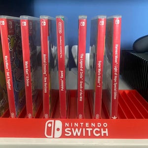 May include: A red Nintendo Switch game display stand with eight slots for game cartridges. The stand has the words "NINTENDO SWITCH" printed on the front. The stand is filled with various Nintendo Switch game cartridges, including "Pok&eacute;mon: Let's Go, Pikachu!", "WarioWare: Get It Together!", "Mario Kart 8 Deluxe", "Animal Crossing: New Horizons", "Pok&eacute;mon Sword", "The Legend of Zelda: Breath of the Wild", "Super Mario Maker 2", and "Harvest Moon: Light of Hope Special Edition".