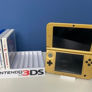 May include: A gold Nintendo 3DS handheld gaming console with a white stand. The console is closed and the top screen is black. The bottom screen is also black and has a white plus button and a white circle button. The console is sitting on a white surface. There are three game cartridges in a white stand that says "Nintendo 3DS" in black text. The game cartridges are for Pokemon Alpha Sapphire, Pokemon X, and Pokemon Y.