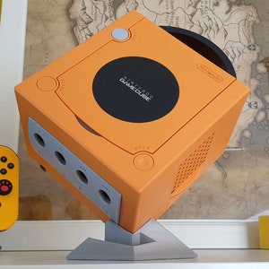 May include: Orange Nintendo GameCube console with a black disc tray and the Nintendo GameCube logo. The console is mounted on a grey stand. The console has four controller ports and a power button. A yellow controller is visible in the background.