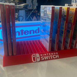 May include: A red and white Nintendo Switch game display stand with a blue and white light-up sign that says "Nintendo". The stand has slots for 10 game cartridges and features the Nintendo Switch logo on the front. The stand is filled with various Nintendo Switch game cartridges, including Animal Crossing: New Horizons, Mario Kart 8 Deluxe, Pok&eacute;mon Sword, Pok&eacute;mon Legends: Arceus, Harvest Moon: Light of Hope Special Edition, WarioWare: Get It Together!, Super Mario Maker 2, The Legend of Zelda: Breath of the Wild, and Pok&eacute;mon Shield.