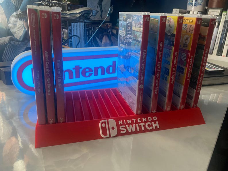 May include: A red and white Nintendo Switch game display stand with a blue and white light-up sign that says "Nintendo". The stand has slots for 10 game cartridges and features the Nintendo Switch logo on the front. The stand is filled with various Nintendo Switch game cartridges, including Animal Crossing: New Horizons, Mario Kart 8 Deluxe, Pok&eacute;mon Sword, Pok&eacute;mon Legends: Arceus, Harvest Moon: Light of Hope Special Edition, WarioWare: Get It Together!, Super Mario Maker 2, The Legend of Zelda: Breath of the Wild, and Pok&eacute;mon Shield.