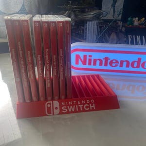 May include: A red Nintendo Switch game storage stand with a white Nintendo logo and the text "NINTENDO SWITCH". The stand is holding a collection of red Nintendo Switch game cartridges. A white and blue light-up sign with the text "Nintendo" is in the background.