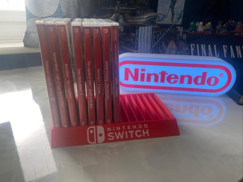 May include: A red Nintendo Switch game storage stand with a white Nintendo logo and the text "NINTENDO SWITCH". The stand is holding a collection of red Nintendo Switch game cartridges. A white and blue light-up sign with the text "Nintendo" is in the background.