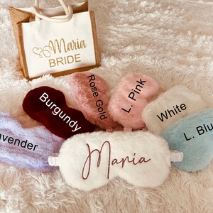 A display of fluffy sleep masks in various colors labeled with names of the shades: Lavender, Burgundy, Rose Gold, L. Pink, White, and L. Blue. A white mask in the center reads "Maria" in rose gold glitter script. A matching "Maria BRIDE" tote bag sits behind the masks on a white fuzzy rug.