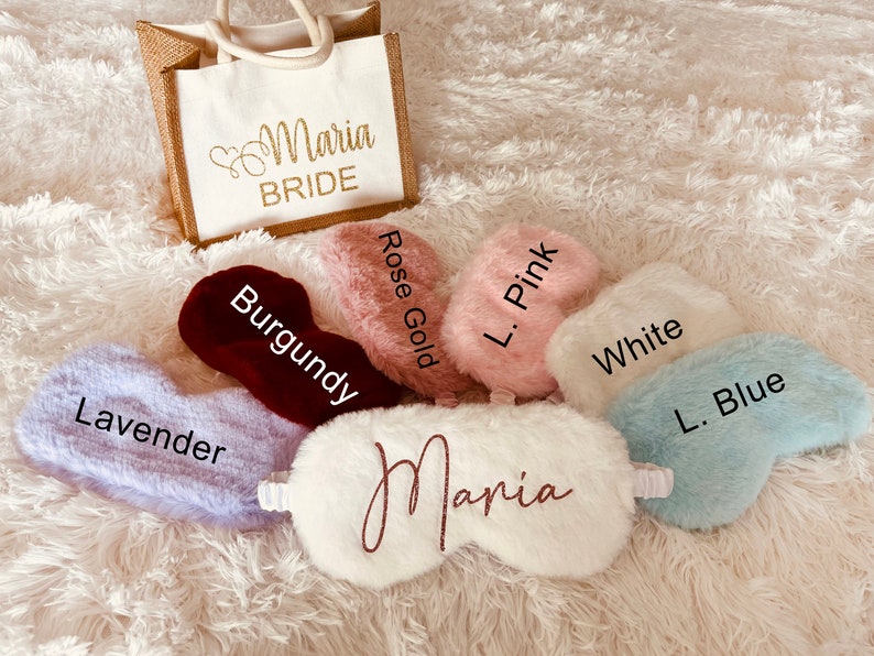 A display of fluffy sleep masks in various colors labeled with names of the shades: Lavender, Burgundy, Rose Gold, L. Pink, White, and L. Blue. A white mask in the center reads "Maria" in rose gold glitter script. A matching "Maria BRIDE" tote bag sits behind the masks on a white fuzzy rug.