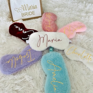 Personalized sleep masks are arranged in a circular layout around a central white mask with the name "Maria." Each mask is in a different color and has a name embroidered in glittery script: Isabella (lavender), Amelia (light blue), Olivia (light pink), Charlotte (rose gold), and Emma (burgundy). A matching white and jute bride tote bag labeled "Maria BRIDE" in gold glitter sits at the top left.