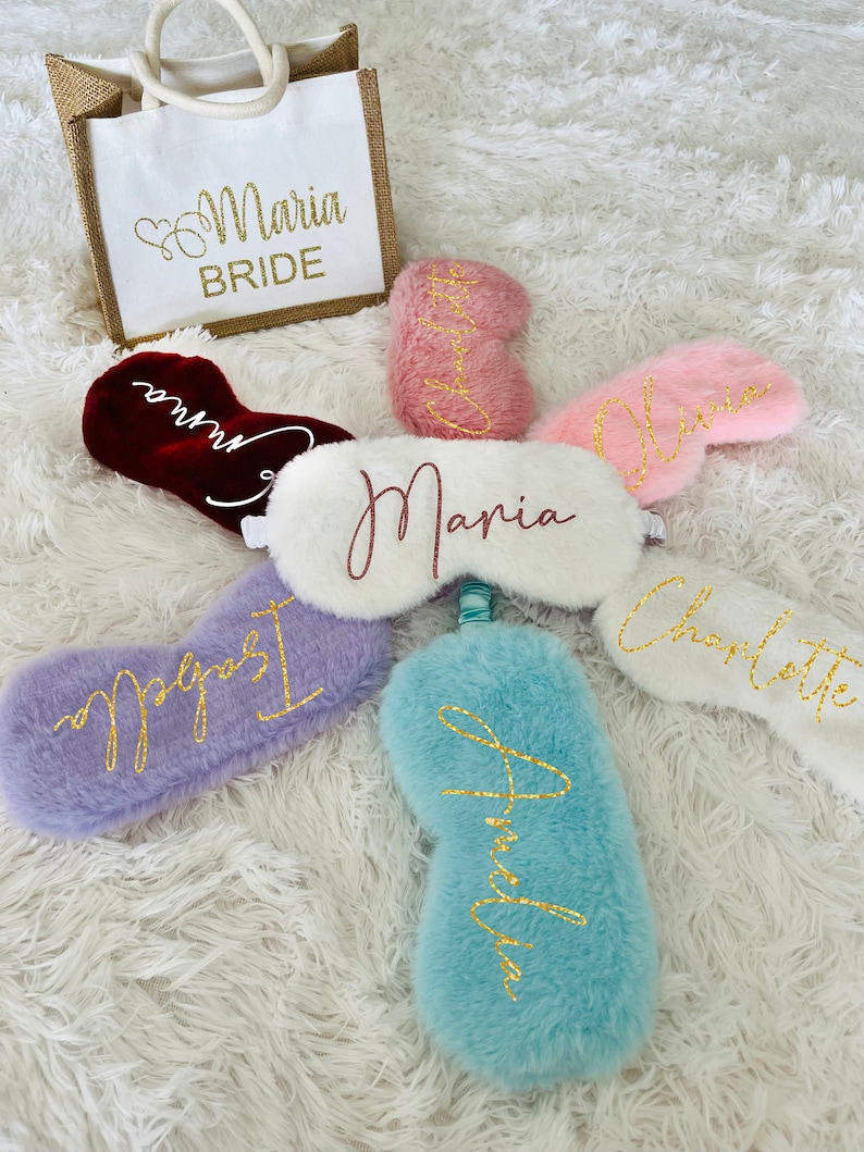 Personalized sleep masks are arranged in a circular layout around a central white mask with the name "Maria." Each mask is in a different color and has a name embroidered in glittery script: Isabella (lavender), Amelia (light blue), Olivia (light pink), Charlotte (rose gold), and Emma (burgundy). A matching white and jute bride tote bag labeled "Maria BRIDE" in gold glitter sits at the top left.