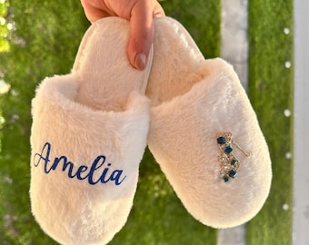Può includere: Ciabatte bianche e soffici con il nome Amelia ricamato su una ciabatta e un ciondolo per scarpe dorato e blu con gioielli sull'altra ciabatta.