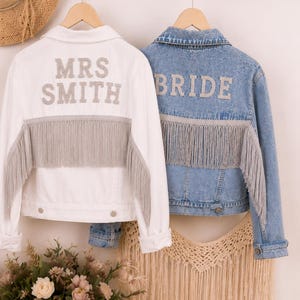 Puede incluir: Dos chaquetas vaqueras cuelgan de una pared. Una es blanca con "MRS SMITH" en letras plateadas y flecos plateados. La otra es de mezclilla azul con "BRIDE" en letras plateadas y flecos plateados. Ropa de boda.