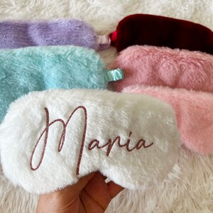 A hand holds a fluffy white personalized sleep mask that reads "Maria" in rose gold glitter script. Several other sleep masks in pastel colors including lavender, light blue, burgundy, and light pink are laid out on a soft white fuzzy surface.