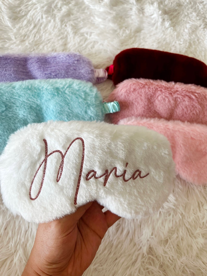 A hand holds a fluffy white personalized sleep mask that reads "Maria" in rose gold glitter script. Several other sleep masks in pastel colors including lavender, light blue, burgundy, and light pink are laid out on a soft white fuzzy surface.