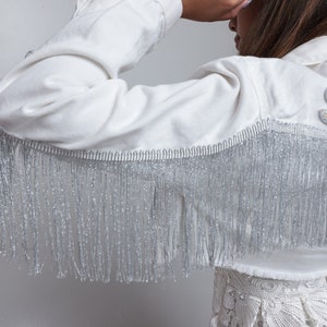 Pode incluir: Jaqueta de ganga branca com acabamento em franjas prateadas e um design "W.S." com pérolas nas costas.