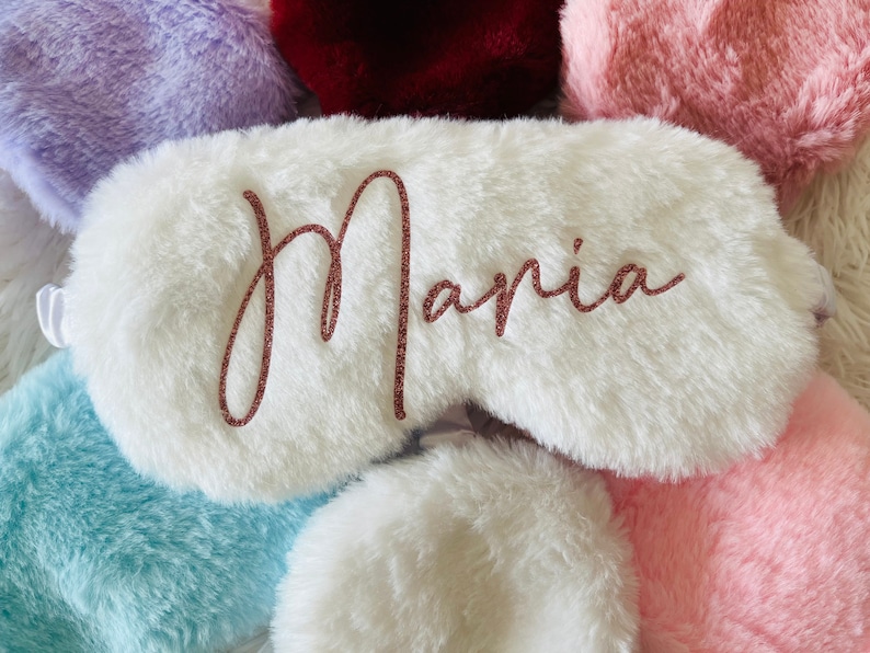 Close-up of a fluffy white sleep mask with the name "Maria" written in rose gold glitter script. Surrounding the mask are other colorful fluffy sleep masks in shades of lavender, burgundy, light pink, light blue, and off-white, arranged on a soft white fur background.