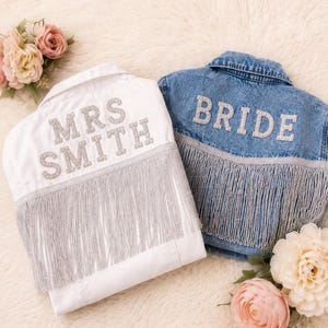 May include: Two denim jackets, one white and one blue, are displayed. The white jacket has "MRS SMITH" in silver letters, and the blue jacket says "BRIDE." Both jackets feature silver fringe along the bottom. Floral arrangements surround the jackets.
