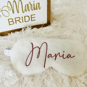 A white fluffy sleep mask with the name "Maria" in rose gold script is placed in front of a white and jute tote bag that says "Maria BRIDE" in glittery gold lettering. Everything is set on a soft white shag rug.