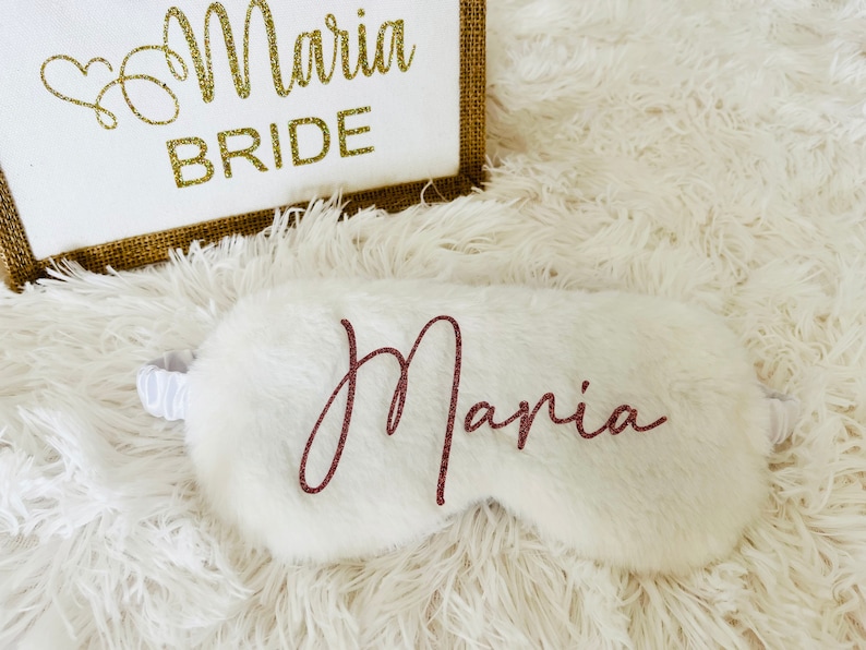 A white fluffy sleep mask with the name "Maria" in rose gold script is placed in front of a white and jute tote bag that says "Maria BRIDE" in glittery gold lettering. Everything is set on a soft white shag rug.