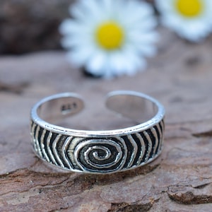 May include: A silver adjustable toe ring featuring a spiral and wave pattern. The ring is open at the top and rests on a brown, textured surface. Two white flowers with yellow centres are blurred in the background.