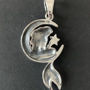 May include: Silver pendant featuring a mermaid sitting on a crescent moon, holding a star. The pendant has a detailed design and a polished finish.