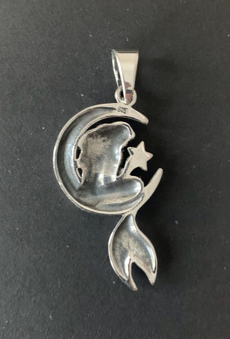 May include: Silver pendant featuring a mermaid sitting on a crescent moon, holding a star. The pendant has a detailed design and a polished finish.