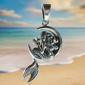 May include: A silver pendant featuring a mermaid sitting on a crescent moon holding a star. The pendant has a detailed design and is perfect for a necklace.