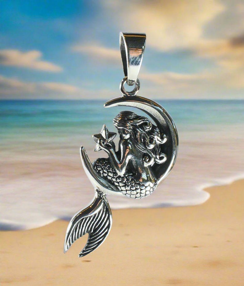 May include: A silver pendant featuring a mermaid sitting on a crescent moon holding a star. The pendant has a detailed design and is perfect for a necklace.