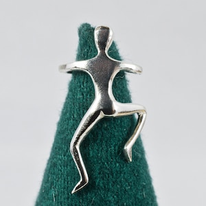Puede incluir: Un anillo plateado con la forma de una figura humana estilizada, con los brazos extendidos y una pierna doblada. El anillo se muestra sobre un fondo verde oscuro texturizado, con un fondo blanco liso.
