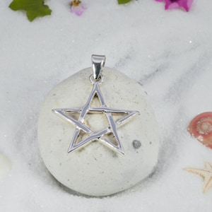 May include: A silver-colored pentagram pendant with a loop for a chain. The star-shaped pendant is displayed on a white stone, with a background of white sand, flowers, and seashells. The pendant is a symbol of faith and spirituality.