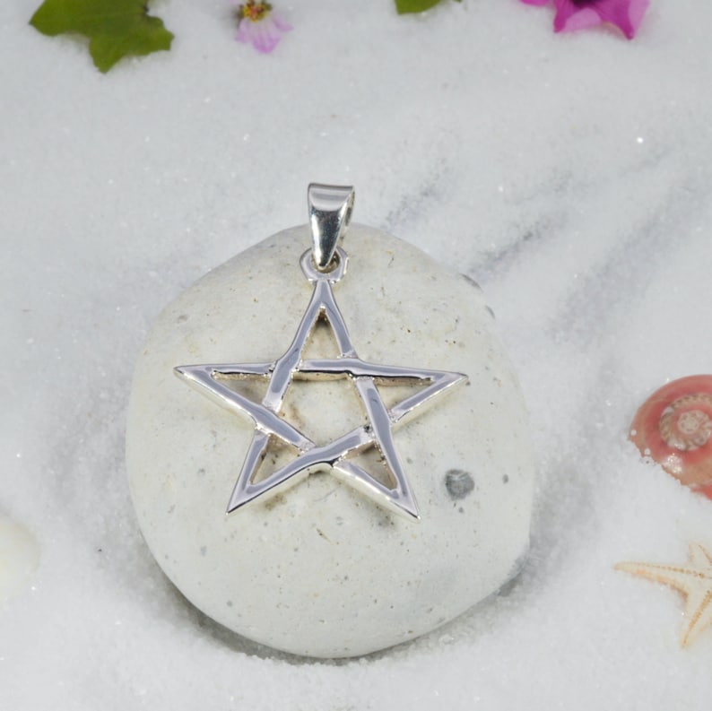 May include: A silver-colored pentagram pendant with a loop for a chain. The star-shaped pendant is displayed on a white stone, with a background of white sand, flowers, and seashells. The pendant is a symbol of faith and spirituality.