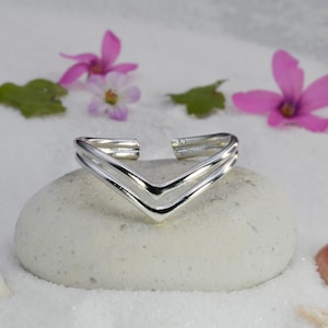May include: A silver V-shaped bracelet with a double-band design, displayed on a smooth, pale stone. The background includes white sand, pink flowers, green leaves, a seashell, and a starfish.