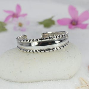 May include: A silver cuff bracelet with a textured design, resting on a white stone. The bracelet has a polished center band and rope-like detailing on the edges. Pink flowers and a seashell are in the background.
