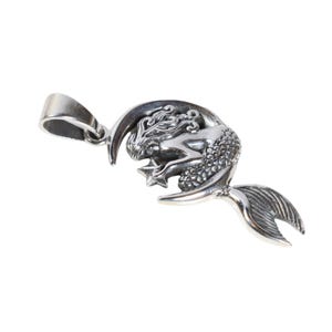 May include: A silver pendant featuring a mermaid curled around a crescent moon, holding a star. The pendant has a loop for attaching to a chain.
