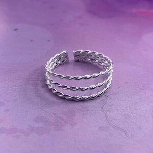 Könnte beinhalten: Ein silberfarbener Ring mit geflochtenem Design. Der Ring hat drei Bänder aus verdrehtem Metall, wodurch ein strukturiertes und kompliziertes Aussehen entsteht. Der Ring ist vor einem weichen, lila Hintergrund platziert.