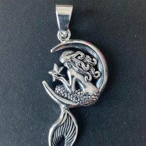 May include: A silver pendant featuring a mermaid holding a star, sitting on a crescent moon. The mermaid's tail extends below the moon.