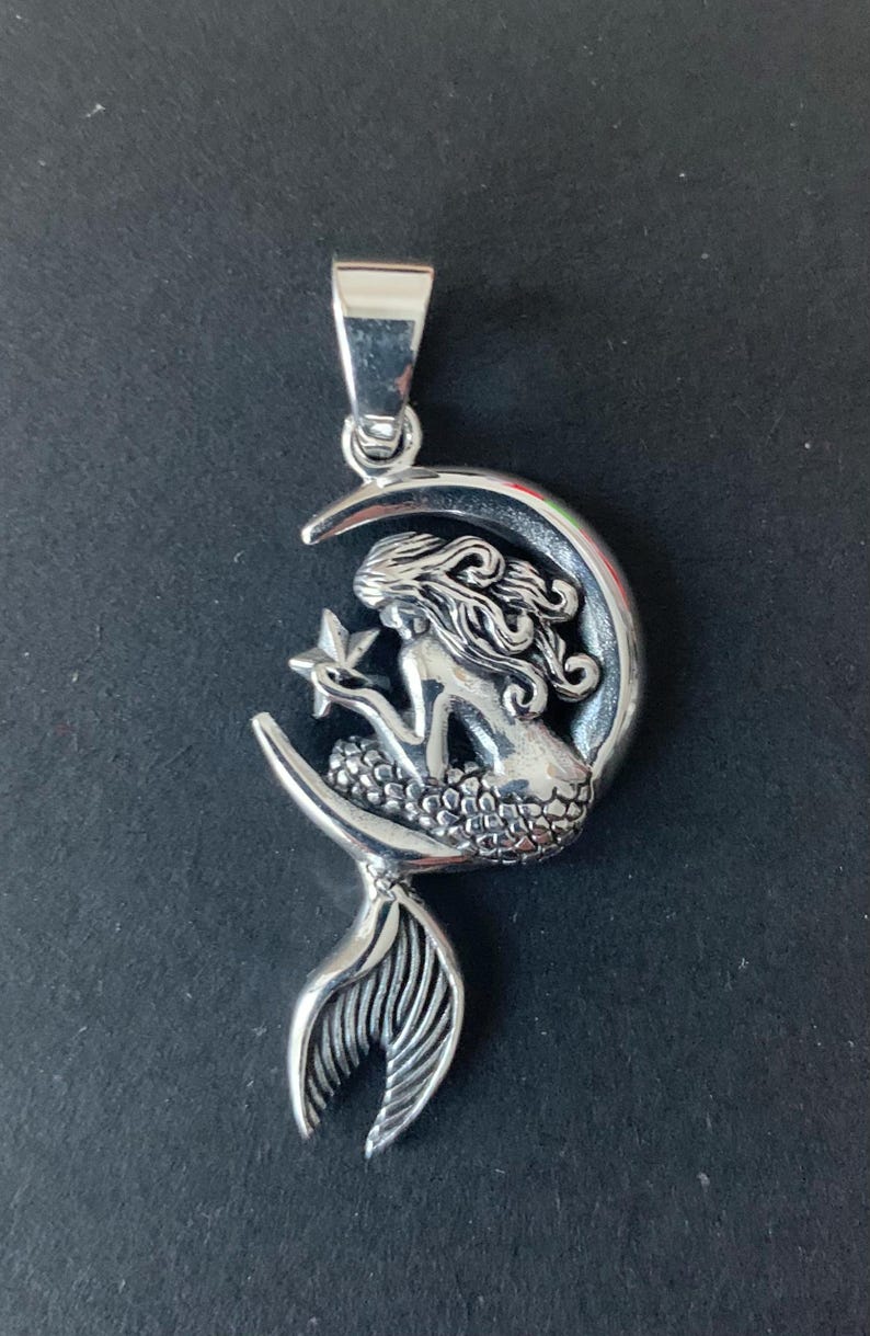 May include: A silver pendant featuring a mermaid holding a star, sitting on a crescent moon. The mermaid's tail extends below the moon.