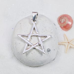May include: A silver-colored pentagram pendant with a loop for a chain. The pendant is displayed on a light gray stone, with a pink seashell and a starfish in the background. The pendant is a five-pointed star.