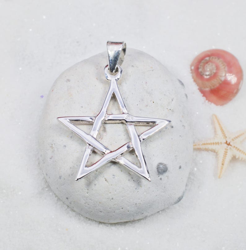 May include: A silver-colored pentagram pendant with a loop for a chain. The pendant is displayed on a light gray stone, with a pink seashell and a starfish in the background. The pendant is a five-pointed star.