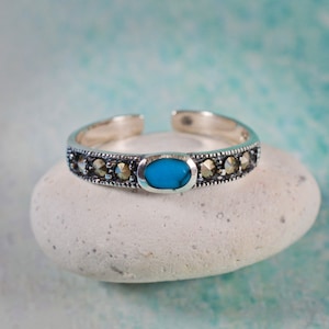 Peut inclure: Une bague d'orteil argentée avec un design ouvert. La bague présente une pierre centrale ovale turquoise et est ornée de petites pierres rondes de couleur foncée. La bague est présentée sur une pierre blanche et arrondie sur fond bleu clair.