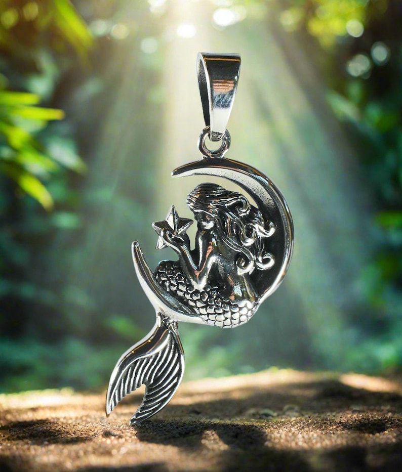 May include: A silver pendant featuring a mermaid sitting on a crescent moon holding a star. The pendant has a detailed design and is perfect for a necklace.