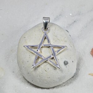 May include: A silver-colored pendant in the shape of a five-pointed star. The pendant is suspended from a small loop at the top. The star is resting on a light-colored stone, set against a background of white sand.