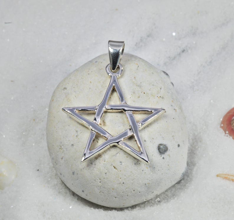 May include: A silver-colored pendant in the shape of a five-pointed star. The pendant is suspended from a small loop at the top. The star is resting on a light-colored stone, set against a background of white sand.