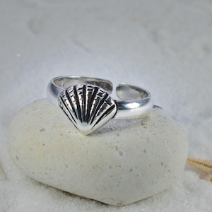 May include: A silver ring featuring a seashell design. The adjustable ring is displayed on a white stone, with a starfish and seashell in the background. The seashell on the ring has detailed, textured lines.