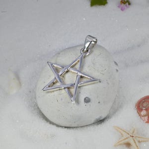 May include: A silver-colored pendant in the shape of a five-pointed star. The pendant is displayed on a light gray stone, set against a backdrop of white sand and small seashells. The star has a loop for a chain.