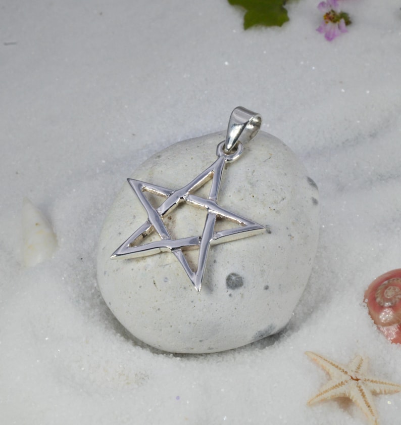 May include: A silver-colored pendant in the shape of a five-pointed star. The pendant is displayed on a light gray stone, set against a backdrop of white sand and small seashells. The star has a loop for a chain.