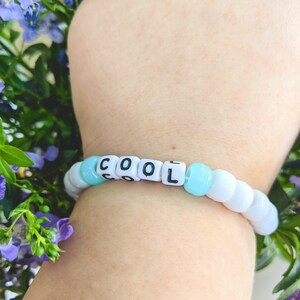 May include: A beaded bracelet with the word "COOL" spelled out in white letter beads. The bracelet features light blue and white beads. The bracelet is worn on a wrist, with a background of purple and green foliage.